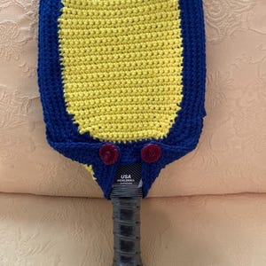 May include: A handmade pickleball paddle cover. The cover is crocheted with a yellow center and a blue border. Two small red flower accents are on the top. The paddle handle is black, and the text "USA Pickleball Approved" is visible.