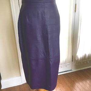 May include: A knee-length, dark purple leather skirt. The skirt is displayed on a mannequin, showcasing its tailored fit and smooth texture. The skirt has a classic design with a high waist and a straight silhouette, suitable for various occasions.