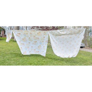 May include: Two large rectangular fabric sheets and one smaller piece hanging on a clothesline. The fabric is white with a repeating pattern of small, light-colored illustrations of children and animals in soft shades of yellow, blue, and brown.