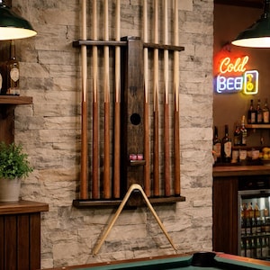 May include: A dark wood wall-mounted pool cue rack holding eight light-colored wooden billiard cues. The rack features a central vertical support with a small shelf holding two pink chalk cubes and a wooden triangle rack hanging below.