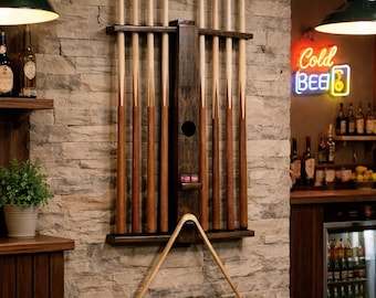 May include: A dark wood wall-mounted pool cue rack holding eight light-colored wooden billiard cues. The rack features a central vertical support with a small shelf holding two pink chalk cubes and a wooden triangle rack hanging below.