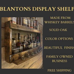 May include: A wooden shelf made from whiskey barrels, with two shelves and a pull-out drawer. The shelf is stained a dark brown color and has a rustic look. It is filled with bottles of liquor, including Blanton's, Evan Williams, and Russell's Reserve. The shelf is labeled "BLANTONS DISPLAY SHELF" and has additional text describing its features: "MADE FROM WHISKEY BARRELS", "SOLID OAK", "COLOR OPTIONS", "BEAUTIFUL FINISH", "FAMILY OWNED BUSINESS", and "FREE SHIPPING".