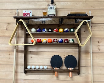 May include: A wooden wall-mounted rack with two shelves for storing pool cues, billiard balls, and table tennis paddles. The top shelf has a rack for 15 billiard balls and two wooden triangle racks. The bottom shelf has a rack for two table tennis paddles. The rack is stained dark brown and has a rustic look.