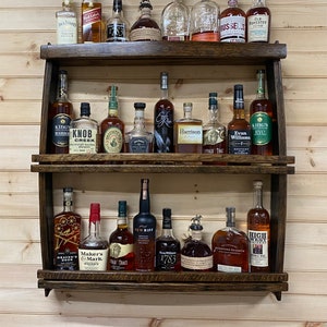 May include: A wooden wall-mounted shelf with two tiers, holding a variety of liquor bottles. The bottles are arranged in rows and include brands like Knob Creek, Evan Williams, Maker's Mark, and High West.