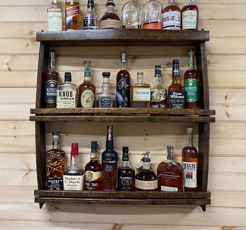 May include: A wooden wall-mounted shelf with two tiers, holding a variety of liquor bottles. The bottles are arranged in rows and include brands like Knob Creek, Evan Williams, Maker's Mark, and High West.