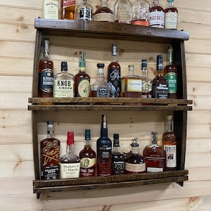 May include: A wooden wall-mounted shelf with two tiers, holding a variety of liquor bottles. The bottles are arranged in rows and are mostly brown glass with labels. Some of the labels include the names "Maker's Mark", "Heaven's Door", "New Riff", "Buffalo Trace", "Evan Williams", and "High West".