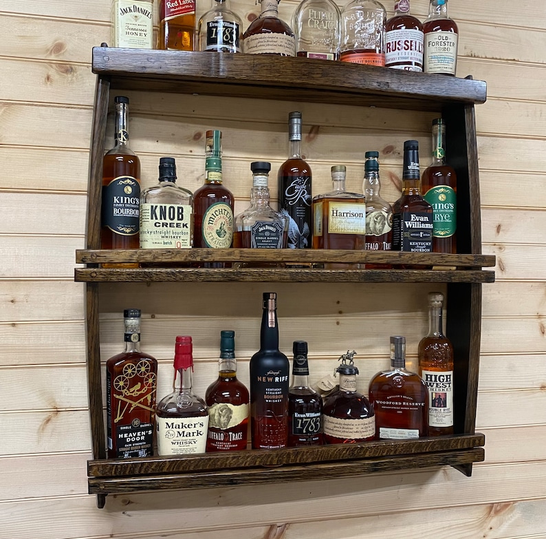 May include: A wooden wall-mounted shelf with two tiers, holding a variety of liquor bottles. The bottles are arranged in rows and are mostly brown glass with labels. Some of the labels include the names "Maker's Mark", "Heaven's Door", "New Riff", "Buffalo Trace", "Evan Williams", and "High West".