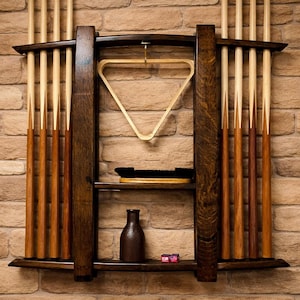 May include: A dark wood wall-mounted pool cue rack featuring two vertical supports, a top shelf holding a light wood triangular ball rack, a middle shelf with a black brush, and a bottom shelf containing a dark bottle and two small red cubes.