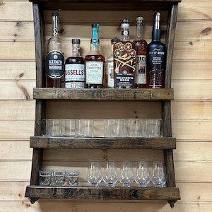 May include: A wooden shelf unit with three shelves, holding various bottles of bourbon whiskey and glasses. The shelf is stained a dark brown color and has a rustic appearance. The bottles are arranged in a staggered pattern, with some bottles on the top shelf and others on the lower shelves. The glasses are arranged in rows on the middle shelf.