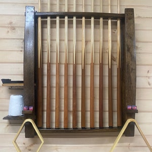 May include: A dark wood wall-mounted pool cue rack with 16 slots for storing cues. The rack has two small shelves on either side for accessories. The rack is mounted on a light wood wall.