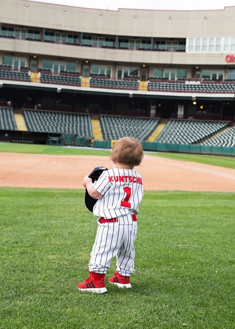 May include: A young child wearing a white and black striped baseball uniform with the name "KUNTSCH" and the number "1" on the back. The child is holding a baseball cap and standing on a green grassy field in front of a baseball stadium.