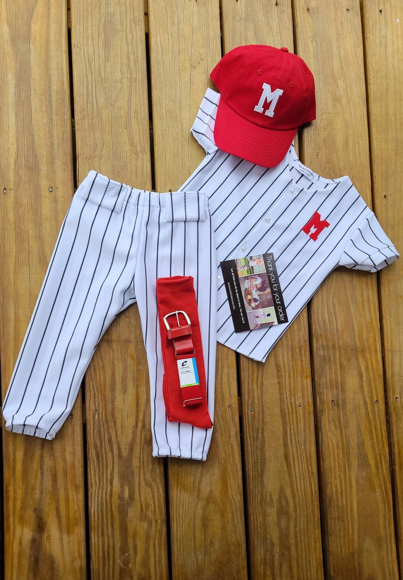 May include: A baseball uniform set for a child, featuring a white and black striped jersey with a red letter "M" on the front, matching pants, a red baseball cap with a white letter "M", and a red belt.