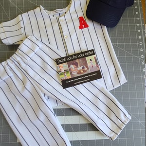 May include: A white and blue striped baseball jersey with a red letter A on the front, white and blue striped pants, and a navy blue baseball cap with a white letter A on the front. The text "Thank you for your order" is on a card with a picture of children.