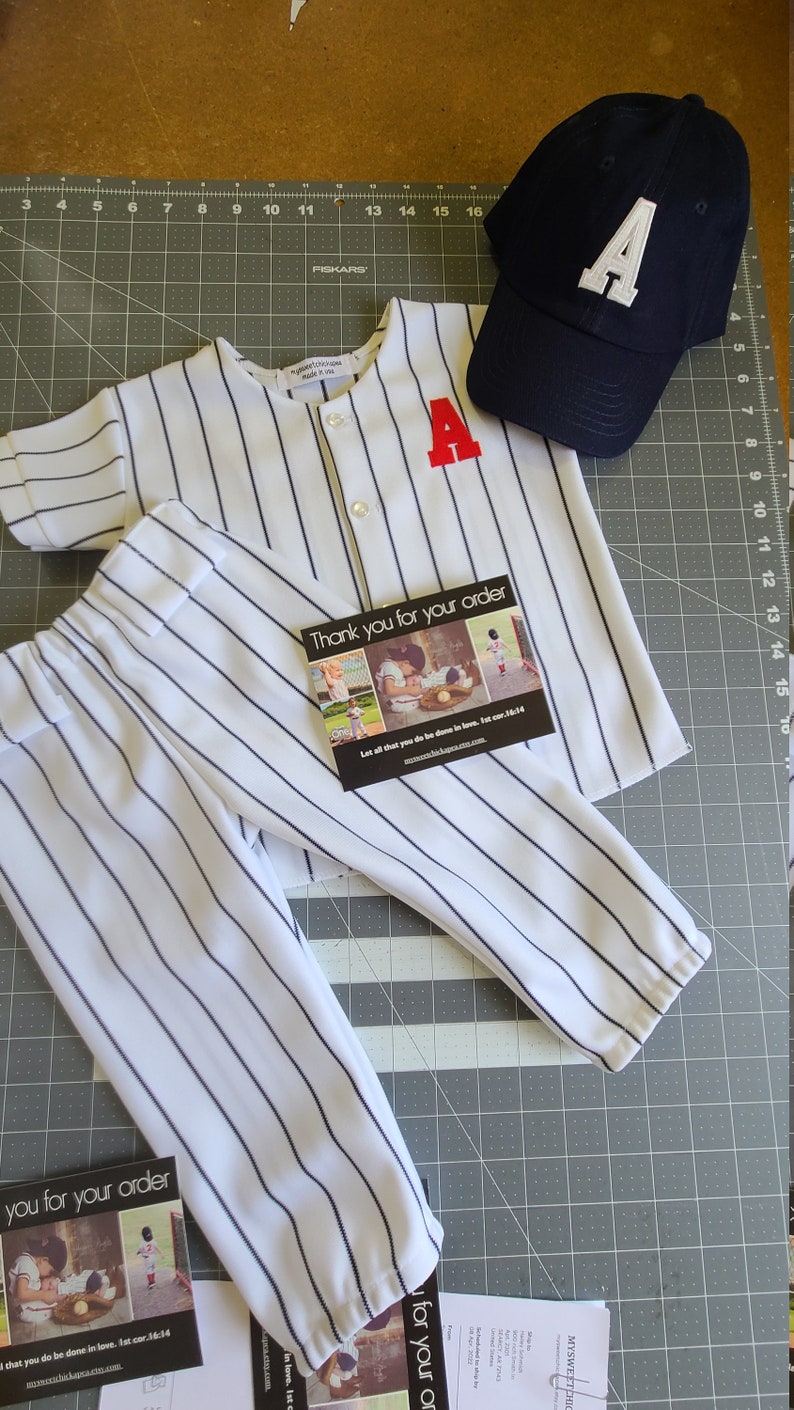 May include: A white and blue striped baseball jersey with a red letter A on the front, white and blue striped pants, and a navy blue baseball cap with a white letter A on the front. The text "Thank you for your order" is on a card with a picture of children.