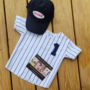 May include: A white and blue striped baseball jersey with a black button and the number one on the front. A black baseball cap with the word "ROOKIE" in red on the front is on top of the jersey. A thank you card with a photo collage is on top of the jersey.