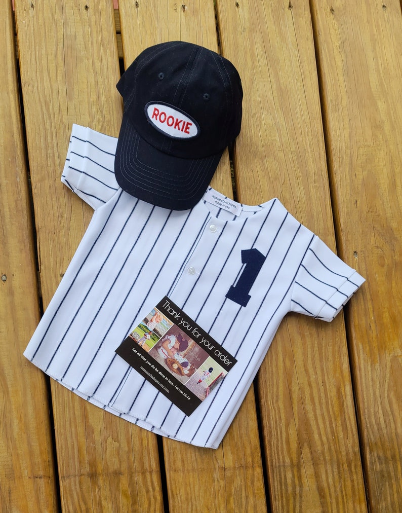 May include: A white and blue striped baseball jersey with a black button and the number one on the front. A black baseball cap with the word "ROOKIE" in red on the front is on top of the jersey. A thank you card with a photo collage is on top of the jersey.