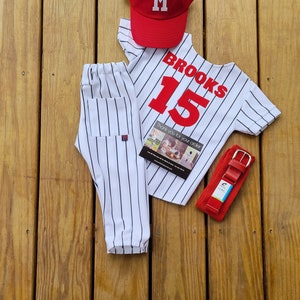 May include: A white and black striped baseball uniform with a red baseball cap, a red belt, and a thank you card. The baseball jersey has the name "BROOKS" and the number "15" on the back.