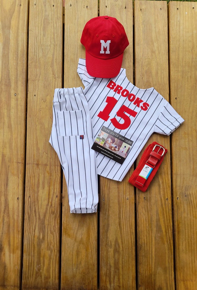 May include: A white and black striped baseball uniform with a red baseball cap, a red belt, and a thank you card. The baseball jersey has the name "BROOKS" and the number "15" on the back.