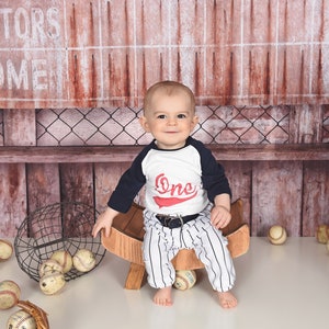 May include: A baby boy wearing a white and navy blue baseball-themed outfit sits on a wooden bench with baseballs scattered around him. The baby is wearing a white shirt with a red "One" on it and navy blue sleeves. The baby is wearing striped pants and a white belt. The baby is smiling and looking at the camera. The background is a wooden wall with a metal sign that says "Visitors Home".