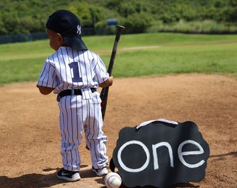 May include: A young child dressed in a baseball uniform holds a bat and stands in front of a black sign that says "one".