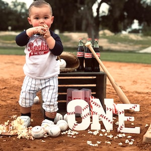 May include: A baby boy wearing a white and blue baseball uniform with the text "Rookie of the Year" is standing on a baseball field. He is holding a piece of cake and there are baseballs and popcorn scattered around him. There is a wooden crate with Coca-Cola bottles and a baseball bat behind him. The word "ONE" is spelled out in large white letters with red stitching.