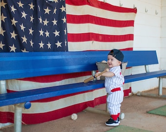May include: A young child wearing a baseball uniform and a black cap is holding a wooden bat and smiling. The child is standing on a blue bench in front of an American flag.