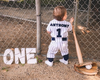 May include: A young child wearing a baseball uniform with the name "Anthony" and the number "1" on the back. The child is standing in front of a chain-link fence with the word "ONE" spelled out in large letters on the ground. A baseball bat and glove are on the ground to the right of the child.