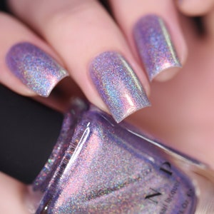 Peut inclure: Gros plan sur un flacon de vernis à ongles violet et des ongles manucurés. Les ongles sont peints avec un vernis holographique qui reflète plusieurs couleurs. Le flacon est tenu dans une main, avec le nom de la marque visible sur l'étiquette. Le flacon de vernis à ongles contient 11,8 ml.