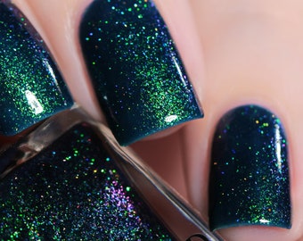 May include: Close-up of dark teal nail polish with green and blue glitter. The polish is applied to fingernails.