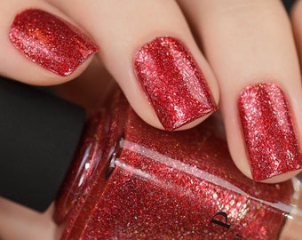 Puede incluir: Una botella de vidrio transparente de esmalte de uñas rojo con purpurina con una tapa negra. La botella está parcialmente cubierta por una mano con esmalte de uñas rojo con purpurina en las uñas.