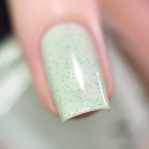 May include: Close-up of a light green nail polish with a subtle shimmer and tiny green glitter particles.