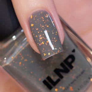 May include: Close-up of a fingernail painted with gray nail polish. The polish has a glittery, speckled effect with orange and gold flakes. The bottle of nail polish is visible in the background, with the brand name "ILNP" printed on it.