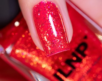 Puede incluir: Primer plano de una uña pintada con esmalte de uñas rojo brillante con purpurina. El esmalte se aplica sobre una base roja, creando un efecto brillante. La uña se sostiene sobre un frasco del mismo esmalte, con el nombre de la marca visible.