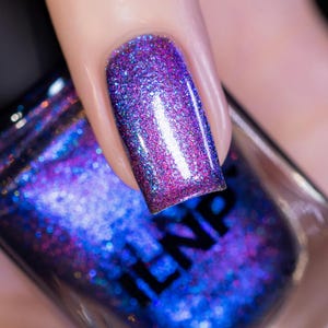 May include: Close-up of a fingernail painted with a vibrant, glittery nail polish. The polish transitions from a deep purple at the base to a shimmering blue at the tip, with iridescent sparkles throughout. The nail is held above a bottle of nail polish.