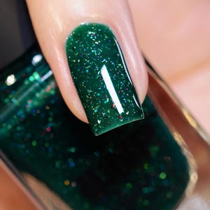 Może przedstawiać: Zbliżenie paznokcia pomalowanego ciemnozielonym lakierem do paznokci. Lakier ma błyszczące wykończenie i jest nakrapiany iryzującym brokatem. Butelka lakieru do paznokci jest widoczna w tle.
