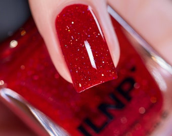 Puede incluir: Primer plano de una botella de esmalte de uñas rojo con un esmalte de uñas rojo con purpurina en un dedo. La botella tiene el nombre de la marca "ILNP" impreso en ella.