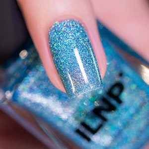 May include: Close-up of a fingernail painted with a sparkling, light blue nail polish. The polish has a glittery, iridescent effect, reflecting various colors. The nail is held over a bottle of matching nail polish, with the brand name "ILNP" visible.