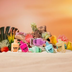 May include: A selection of ILNP Studio Color nail polish bottles in shades of orange, pink, purple, green, and blue. The bottles are displayed on a sandy surface, with flowers and greenery in the background.