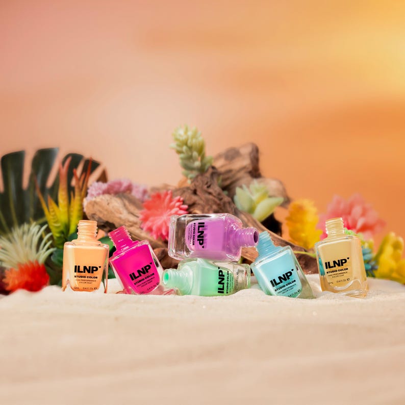 May include: A selection of ILNP Studio Color nail polish bottles in shades of orange, pink, purple, green, and blue. The bottles are displayed on a sandy surface, with flowers and greenery in the background.