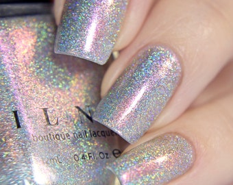 Puede incluir: Una botella de vidrio transparente de esmalte de uñas con un acabado de brillo plateado iridiscente. La botella está etiquetada como "ILNP boutique nail lacquer" y "0.4 fl. oz.". La botella se muestra con una mano que lleva el esmalte de uñas, que es un acabado de brillo plateado iridiscente.