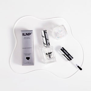 May include: A clear glass bottle of ILNP Glass Candy Ultra Glossy Top Coat nail polish with a black and silver label. The bottle is sitting on a white surface with a black and silver cap and a brush.
