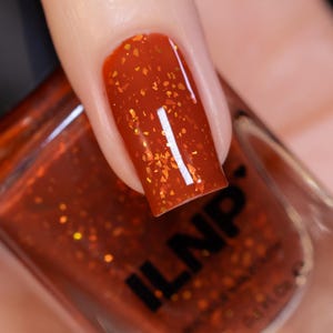 May include: Close-up of a fingernail painted with a vibrant orange nail polish. The polish has a glossy finish with gold glitter flakes. The nail polish bottle is visible in the background, with the brand name "ILNP" printed on it.