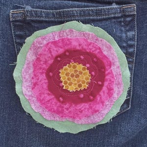 May include: Close-up of a denim jean pocket with a large, layered fabric flower applique. The flower features shades of pink, green, and yellow, with a honeycomb-like center. The denim is a dark blue wash.