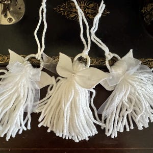 May include: Three handmade white angel ornaments. Each angel features a yarn body, fabric wings, and a loop for hanging. The angels are decorated with sheer ribbon and a small embellishment on the center angel.