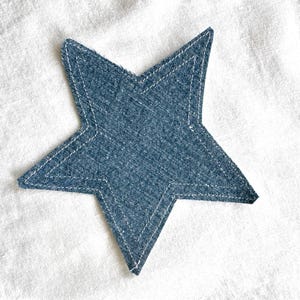 May include: A denim star-shaped patch with white stitching. The star is a medium blue color and is set against a white fabric background. The patch has a five-point design and is likely for decorative purposes.