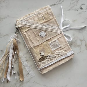 May include: A beige journal with a patchwork design, adorned with lace and a decorative button. A white ribbon wraps around the journal, and a tassel with beads hangs from the side. The journal's cover is made of various fabric pieces.