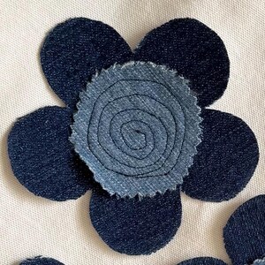 May include: A fabric flower applique featuring five dark navy blue denim petals surrounding a central circular piece of lighter blue denim. The center is detailed with a dark blue spiral stitch pattern and has a frayed, textured edge.