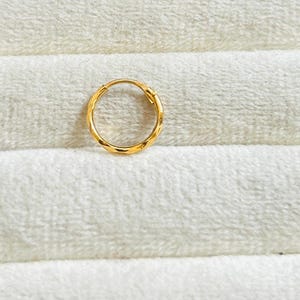 Op de afbeelding: Een enkele gouden oorring met een getextureerd, facetontwerp. De oorring is cirkelvormig en lijkt gemaakt van een glanzend, geel metaal. De oorring is geplaatst tegen een witte, getextureerde achtergrond.