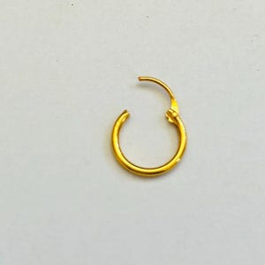 Könnte beinhalten: Ein einzelner goldener Creolen-Ohrring, offen und zum Verschließen bereit. Der Ohrring hat eine helle, glänzende Goldfarbe und ist vor einem schlichten weißen Hintergrund platziert. Der Ohrring ist ein schlichtes, klassisches Design.