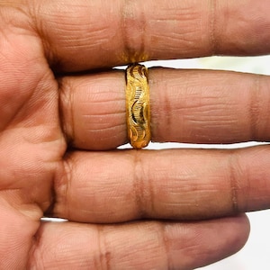 Puede incluir: Un anillo de oro con un patrón intrincado y repetitivo. El anillo es de un color dorado brillante y parece estar hecho de metal. El anillo está en un dedo y los detalles del diseño del anillo son visibles.